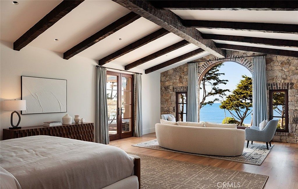 This spacious primary bedroom features a vaulted ceiling with exposed dark wood beams and a prominent stone wall surrounding a large arched window that frames a scenic ocean view. The room is furnished with a neutral-toned bed, a curved sofa, and a long wooden console table, all set against light wood flooring and soft blue curtains. The overall aesthetic is a blend of rustic stone elements and refined, airy coastal design.