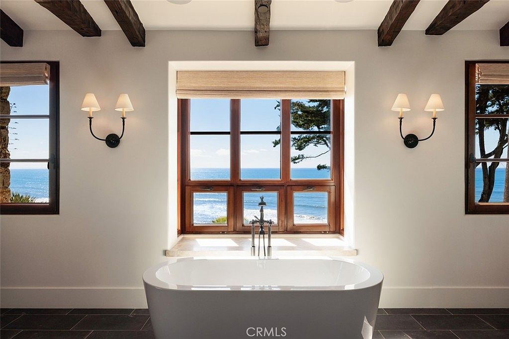 This luxurious primary bathroom features a freestanding white soaking tub positioned directly in front of a large, multi-pane window that frames a stunning ocean view. The room is accented by dark wood ceiling beams, wall-mounted sconces, and a sleek, modern faucet fixture. The neutral walls and dark tile flooring create a serene, high-end atmosphere perfect for a coastal retreat.