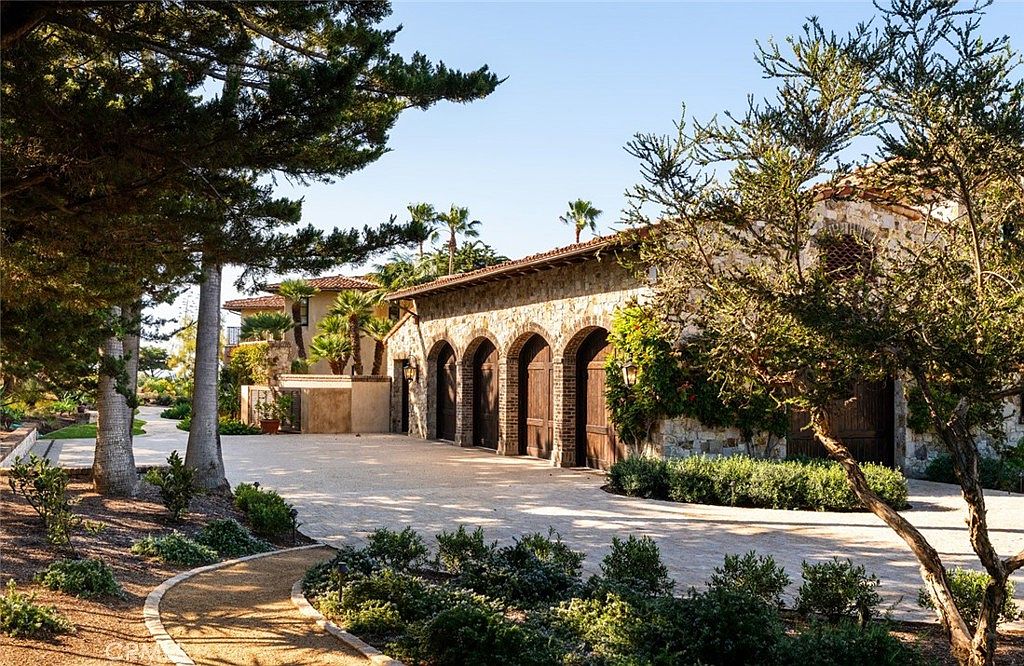 This image captures a Mediterranean-style estate featuring a multi-bay garage with arched wooden doors and stone masonry walls. A wide, light-colored stone driveway leads up to the structure, flanked by manicured landscaping and mature trees. The perspective is a wide-angle shot from the driveway, emphasizing the architectural character and the expansive, sun-drenched outdoor space.