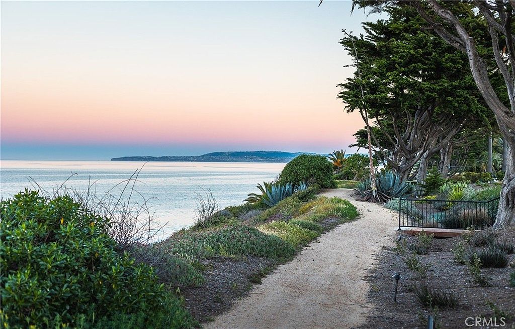 This scenic outdoor pathway winds along a coastal cliffside, offering expansive views of the ocean and a distant headland under a soft, twilight sky. The path is bordered by lush, drought-tolerant landscaping, including agave plants and mature, wind-swept trees that frame the horizon. A simple metal railing marks the edge of the property, creating a serene and picturesque transition between the garden and the natural coastline.