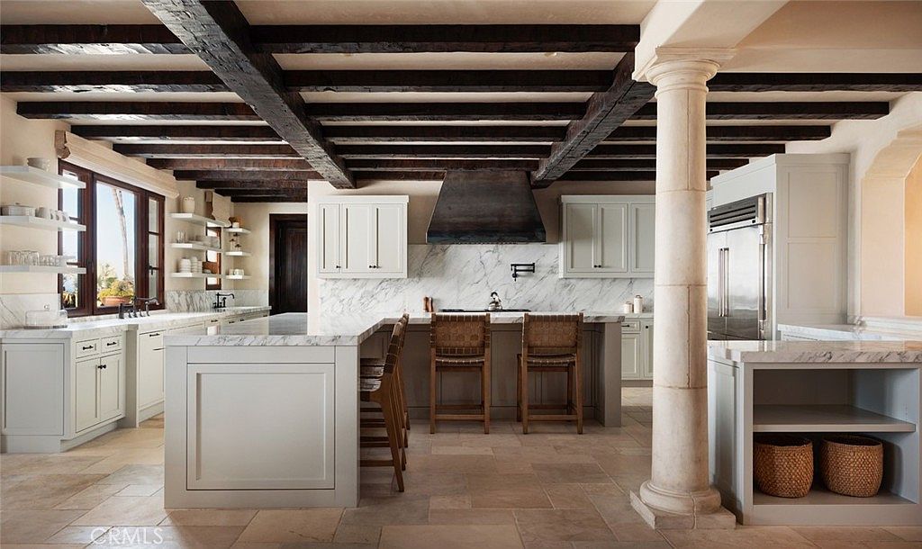 This spacious kitchen features a rustic aesthetic with exposed dark wood ceiling beams and light-toned cabinetry. A large central island with stone countertops provides seating, complemented by a matching stone backsplash and a prominent metal range hood. The room is anchored by a classic stone column and features stone tile flooring, creating a warm and sophisticated atmosphere.