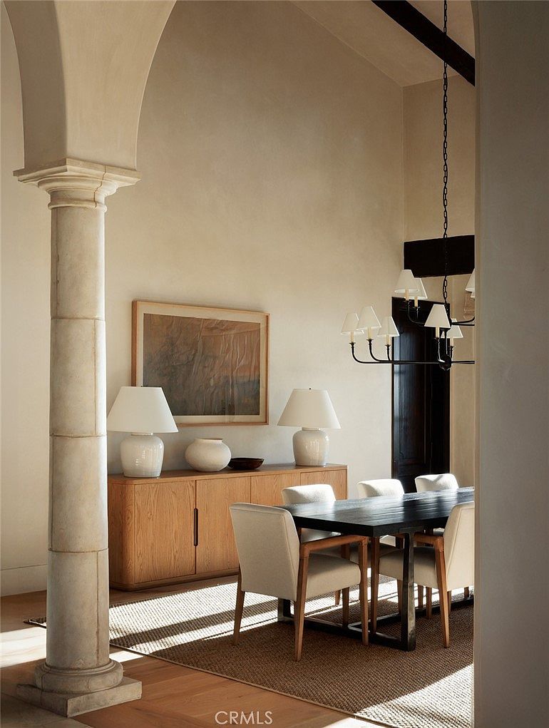 This elegant dining room features a dark wood table surrounded by light-colored upholstered chairs, set against a backdrop of a light wood sideboard topped with two matching table lamps. A large, abstract framed artwork hangs on the neutral wall, while a dramatic black chandelier hangs above the table, adding a modern contrast to the space. The room is framed by a classic stone column in the foreground, creating a sophisticated and airy atmosphere with warm, natural light filtering in.