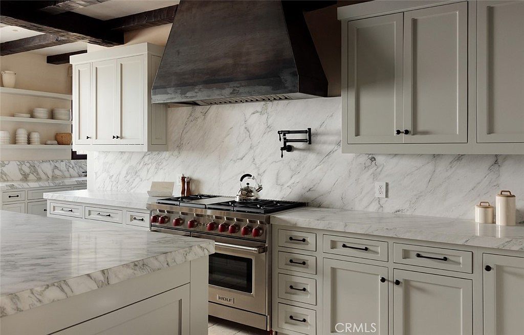 This elegant kitchen features light-colored cabinetry, a professional-grade range with a prominent metal hood, and extensive stone countertops with a distinct veined pattern. A wall-mounted pot filler is centered above the range, set against a matching stone backsplash that extends across the wall. The perspective captures the workspace from a slightly elevated angle, highlighting the clean lines and sophisticated, neutral aesthetic of the culinary space.