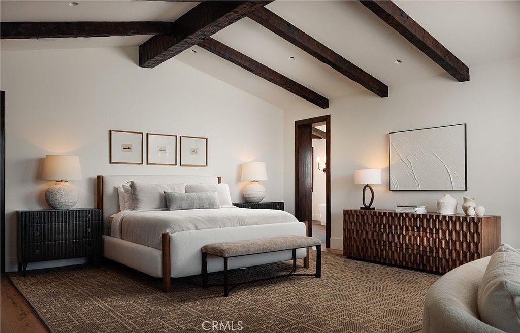This elegant primary bedroom features a vaulted ceiling with exposed dark wood beams, creating a sophisticated and airy atmosphere. The room is anchored by a plush bed with a neutral upholstered headboard, flanked by matching nightstands and textured table lamps. A unique, carved-wood dresser adds visual interest to the space, while a neutral area rug and a bench at the foot of the bed complete the refined, contemporary design.