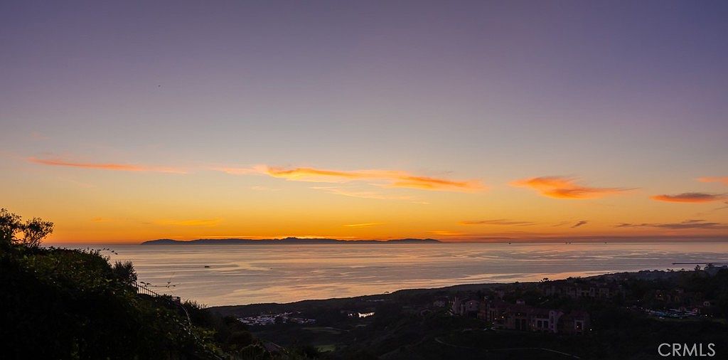 This aerial view captures a stunning sunset over the ocean, with a coastline visible in the distance. The sky is a blend of purple and orange hues, reflecting on the water's surface. In the foreground, there are residential buildings nestled among lush greenery, suggesting a high-end coastal community.