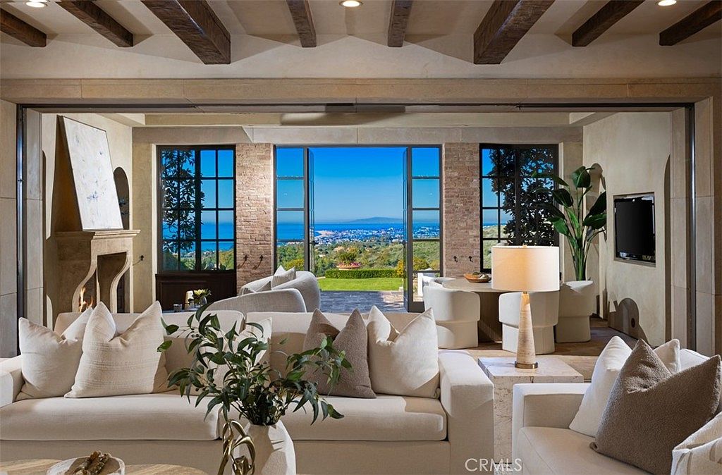 This is an interior shot of a luxurious living room featuring a neutral color palette with white sofas and textured pillows. The room has exposed wooden beams on the ceiling and large windows offering a stunning view of the ocean and landscape. A fireplace and a modern lamp add to the room's sophisticated ambiance.