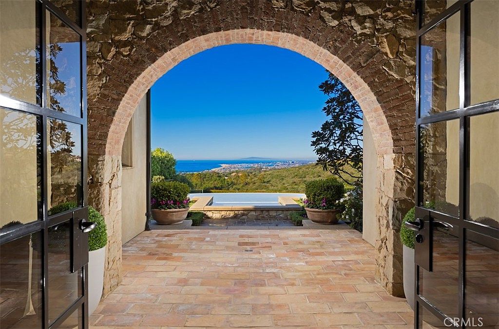 This image showcases a stunning outdoor patio area framed by a rustic stone archway and elegant glass doors. The patio features brick flooring, leading to a view of a pool, lush greenery, and a distant cityscape under a clear blue sky. The scene evokes a sense of luxury and tranquility, highlighting the property's exceptional outdoor living space.