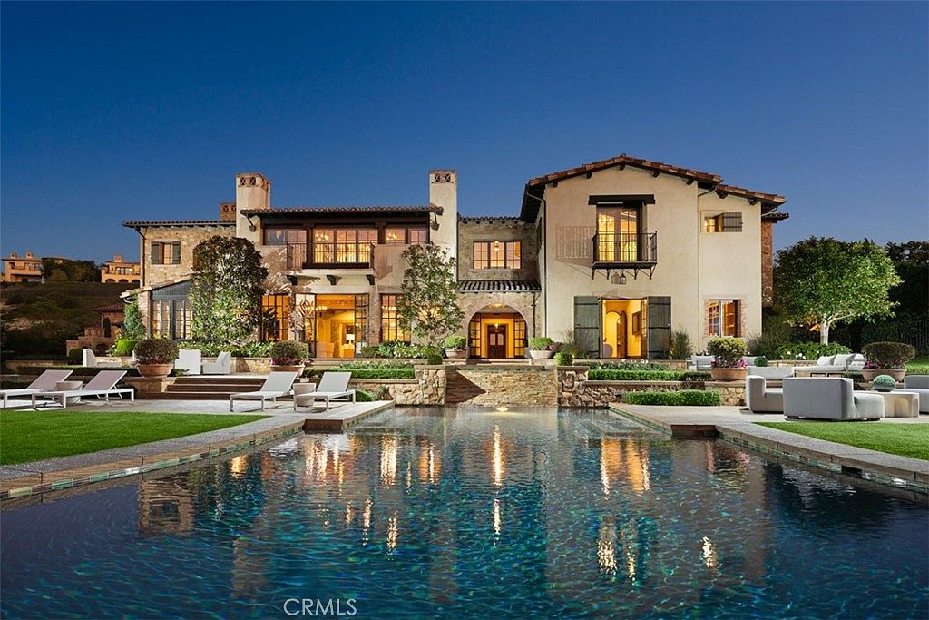 This is a stunning rear exterior view of a luxurious Mediterranean-style home, showcasing a large swimming pool with shimmering reflections. The meticulously landscaped backyard features lounge chairs, outdoor seating, and lush greenery, creating an inviting and serene atmosphere. The architecture of the home includes multiple balconies, arched entryways, and warm lighting, enhancing its elegant appeal.