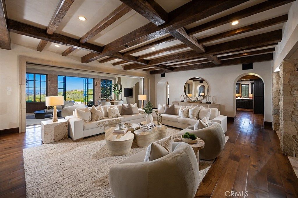 This is an interior shot of a luxurious living room featuring exposed wooden beams on the ceiling, two white sofas, and two armchairs arranged around a set of coffee tables on a large area rug. The room is well-lit with natural light streaming in from large windows, and the hardwood flooring adds warmth to the space. The overall impression is one of comfort and sophistication.