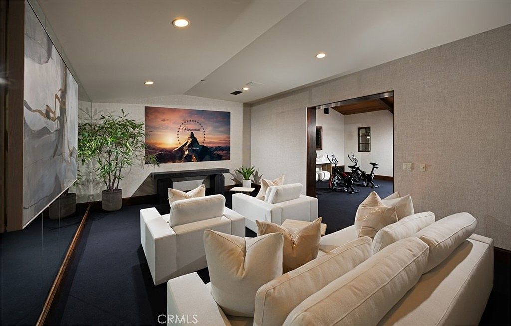 This interior shot showcases a modern living room with a home theater setup. The room features a large screen, comfortable white seating, and a Paramount Pictures logo displayed on the wall. An adjacent room with exercise bikes suggests a home gym, creating a versatile and luxurious space.