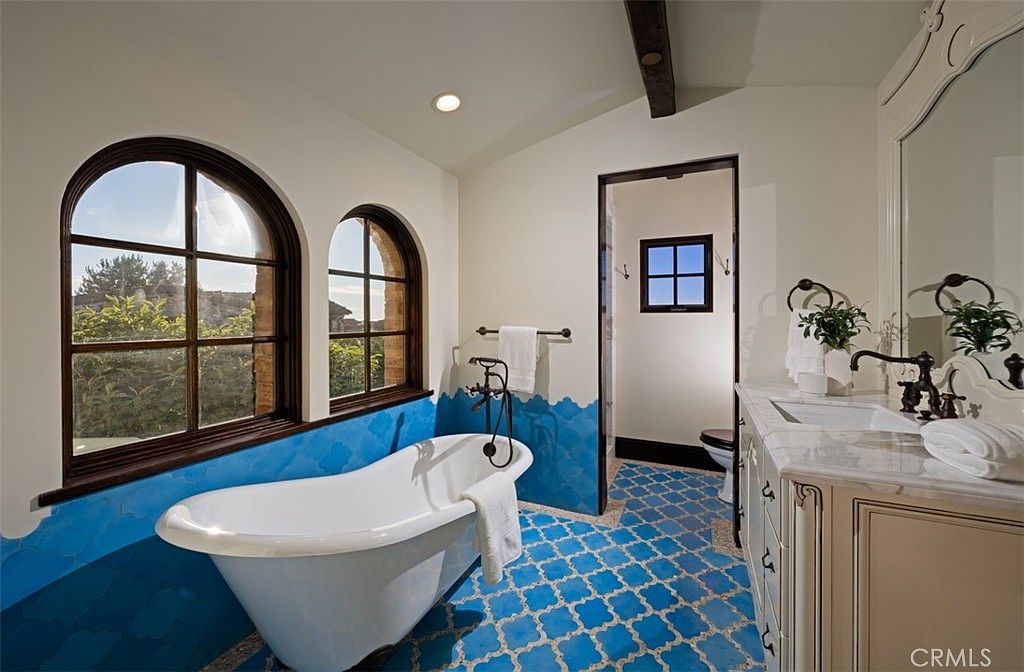 This primary bathroom features a freestanding bathtub positioned beneath arched windows that offer natural light and views. The room is decorated with blue patterned tiles on the floor and lower wall, creating a unique and stylish design. A vanity with a marble countertop and ornate mirror adds a touch of elegance to the space.