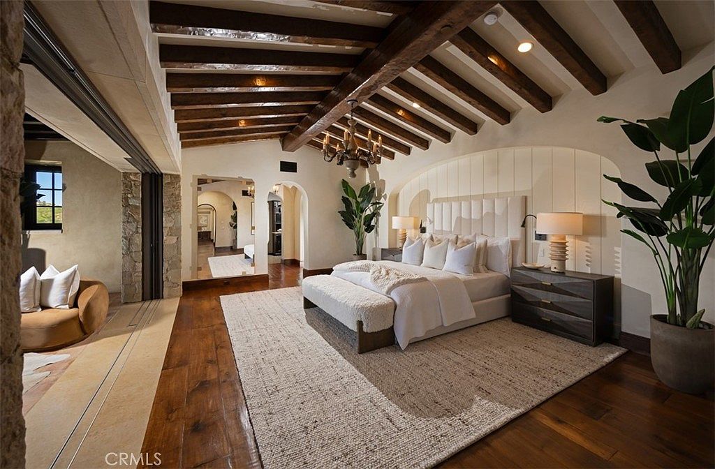 This is a luxurious primary bedroom featuring a king-size bed with a tufted headboard, a neutral-toned rug, and exposed wooden beams on the ceiling. The room has an open layout with a sitting area visible through an arched doorway, and is decorated with large potted plants, creating a serene and inviting atmosphere. The dark wood flooring contrasts with the light walls and bedding, adding depth and warmth to the space.