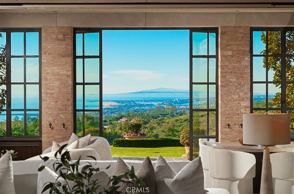 This interior shot showcases a luxurious living room with a stunning view. Large windows frame a picturesque landscape of rolling hills and the ocean, creating a seamless connection between indoor and outdoor spaces. The room is furnished with elegant white sofas and chairs, complemented by a stylish lamp, creating a bright and inviting atmosphere.