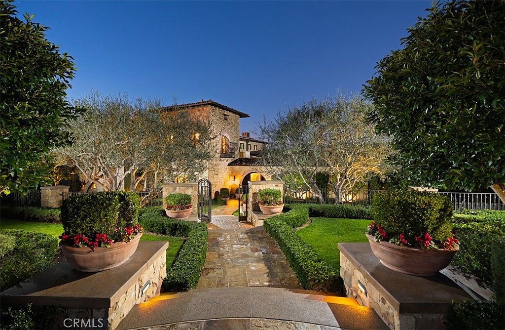 This image showcases a beautifully landscaped yard and garden leading up to a grand estate. The stone pathway is flanked by manicured hedges and potted plants, creating a formal yet inviting entrance. Mature trees and subtle lighting enhance the property's curb appeal, suggesting a luxurious and well-maintained residence.