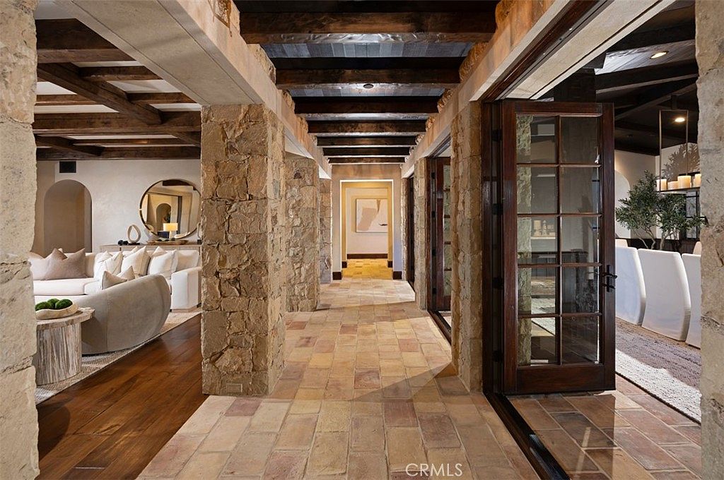 This interior shot showcases a hallway with rustic stone pillars and brick flooring, leading to various rooms. The ceiling features exposed wooden beams, adding to the architectural detail. The open doorways offer glimpses into a living room and dining area, creating a sense of spaciousness and flow.