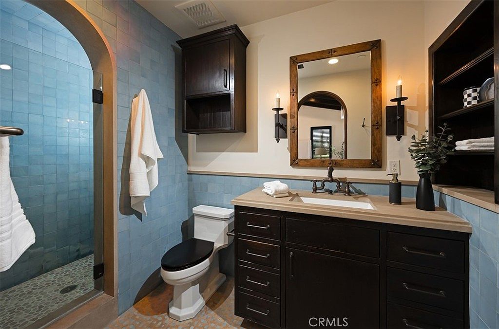 This bathroom features blue tiled walls, a dark wood vanity with a light countertop, and a framed mirror with sconces. A toilet is positioned next to a built-in cabinet, and a glass-enclosed shower is visible to the left. The overall design creates a cozy and stylish space.