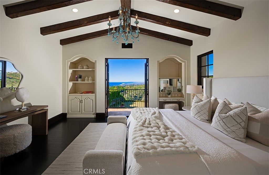 This is a primary bedroom featuring a luxurious bed with multiple pillows and a textured throw blanket. The room has dark wood floors, exposed wood beams on the ceiling, and built-in shelving units on either side of a balcony door that offers a stunning ocean view. A chandelier hangs from the ceiling, adding an elegant touch.