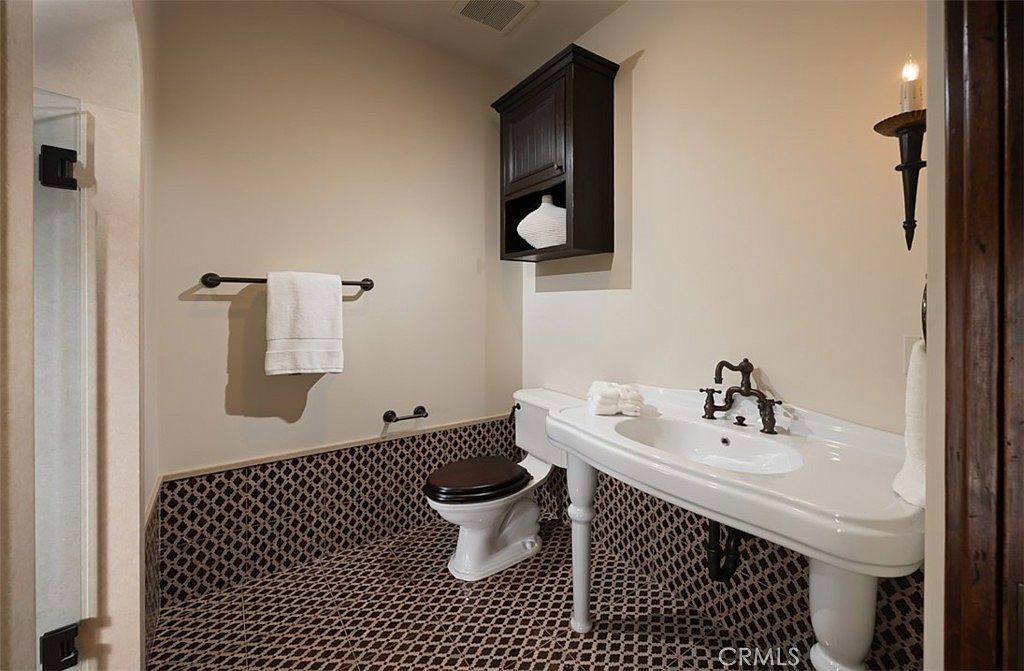 This is a guest bathroom featuring a unique design with dark brown and beige patterned tile flooring and wainscoting. The white pedestal sink with vintage-style faucet and dark wood toilet seat add to the room's character. A dark wood cabinet hangs above the toilet, and a sconce provides warm lighting.