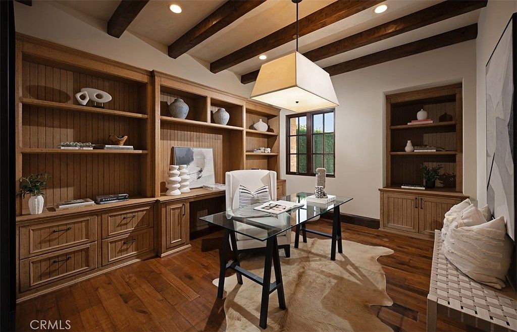 This is an interior shot of a home office or study, featuring custom built-in wooden shelving and cabinetry. A glass-topped desk with black trestle legs sits in the center of the room, illuminated by a modern square pendant light. The room has hardwood floors covered by a hide rug, and a bench with pillows is visible on the right, creating a warm and inviting workspace.