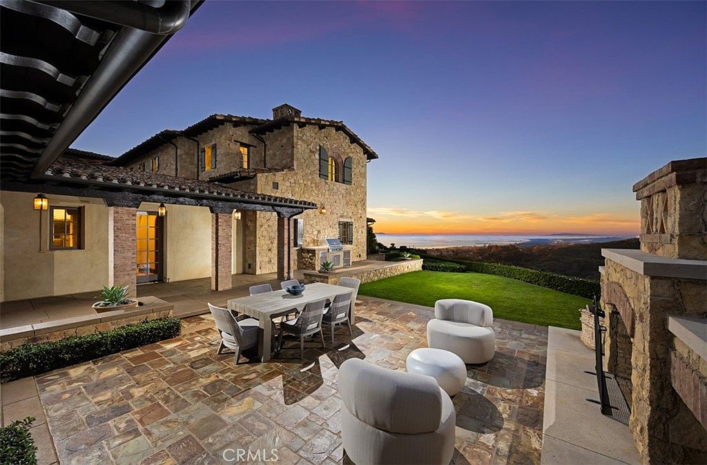 This image showcases a luxurious outdoor patio area at dusk, featuring a stone-paved surface, a dining table with seating, and comfortable lounge chairs. The patio is adjacent to a well-manicured lawn and offers stunning views of the ocean and sunset. The stone facade of the house and outdoor fireplace add to the elegant and inviting atmosphere.