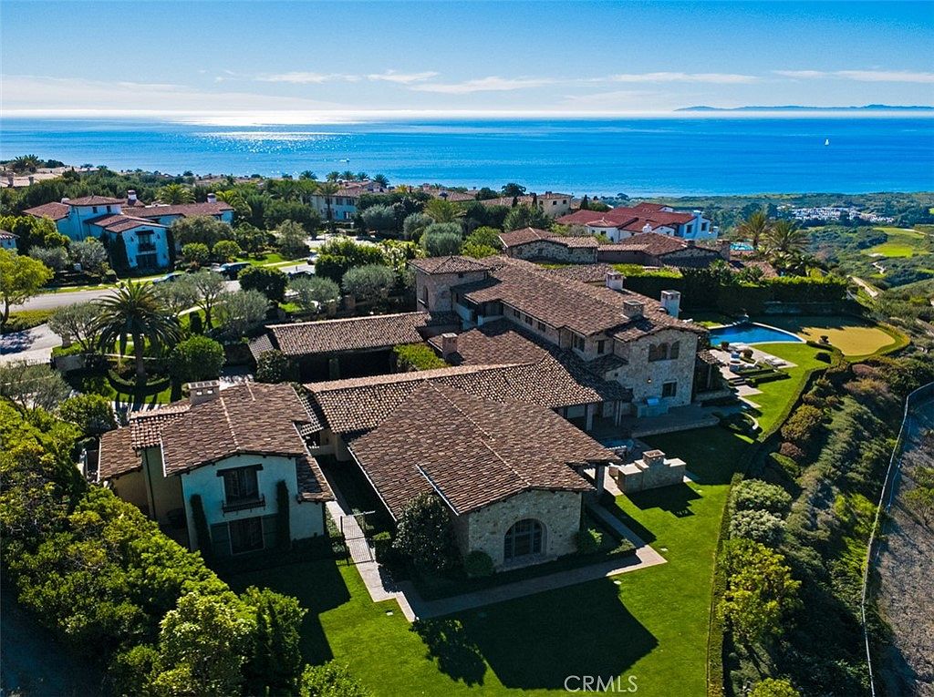 This aerial shot showcases a sprawling estate with a terracotta tile roof, lush green lawns, and mature landscaping. A sparkling blue pool and golf course are visible in the backyard, with a stunning ocean view in the background. The overall impression is one of luxury and privacy.
