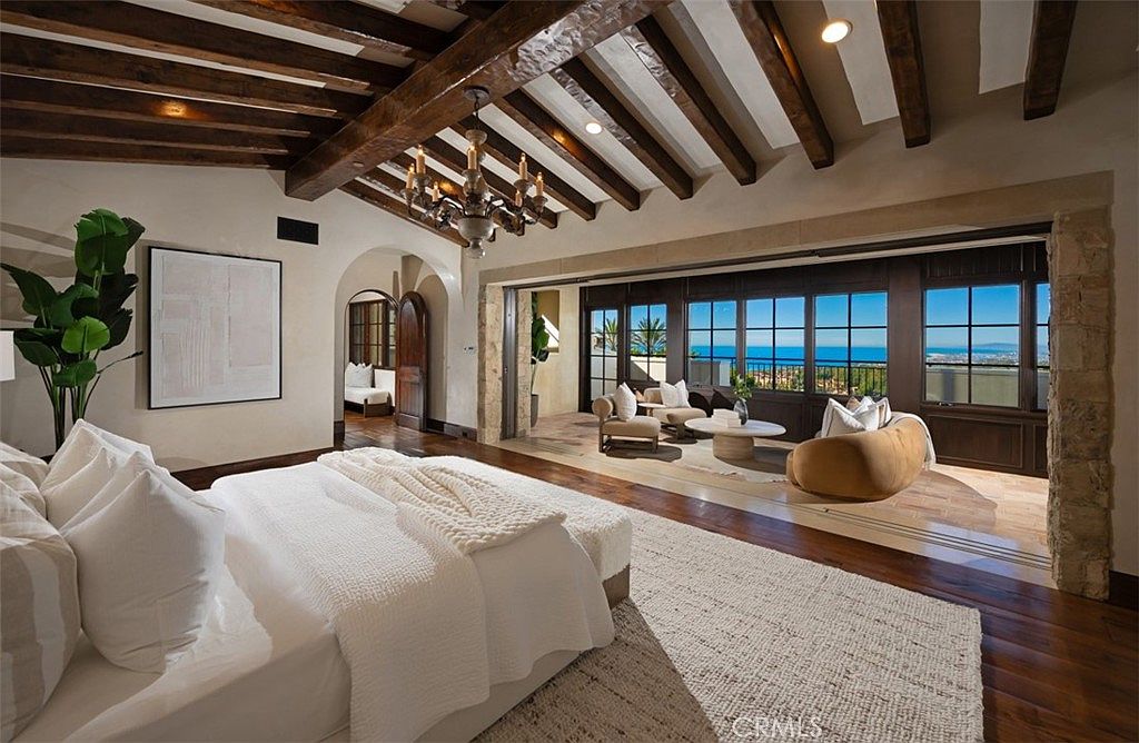 This is a luxurious primary bedroom featuring a king-size bed with white linens, a textured rug, and exposed wooden beams on the ceiling. The room opens to a sitting area with ocean views through large windows, creating a seamless indoor-outdoor living experience. An arched doorway leads to another room, adding to the spacious and elegant feel of the suite.