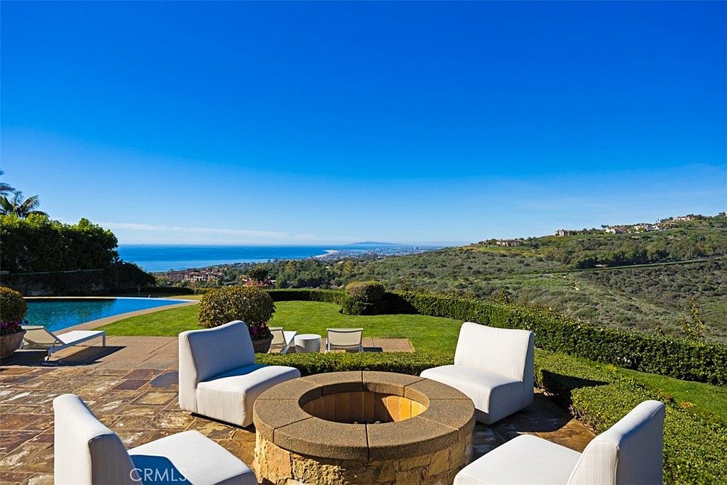 This image showcases a luxurious outdoor patio area with a stunning view. The focal point is a stone fire pit surrounded by modern white chairs, creating an inviting space for relaxation and entertainment. Beyond the patio, a well-manicured lawn leads to a pool and panoramic ocean views, enhancing the property's appeal.