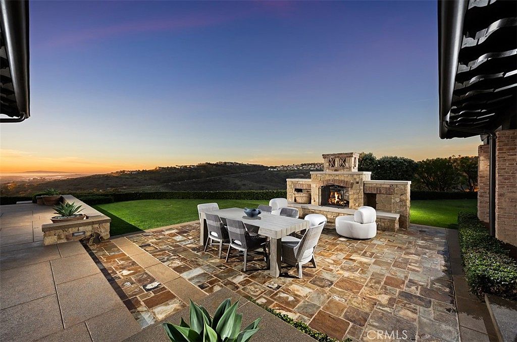 This image showcases a beautifully designed outdoor patio area, perfect for entertaining or relaxing. The space features a stone fireplace, a large dining table with seating, and a comfortable lounge chair. The patio is paved with decorative stones and overlooks a well-maintained lawn and scenic landscape, creating a luxurious and inviting atmosphere.