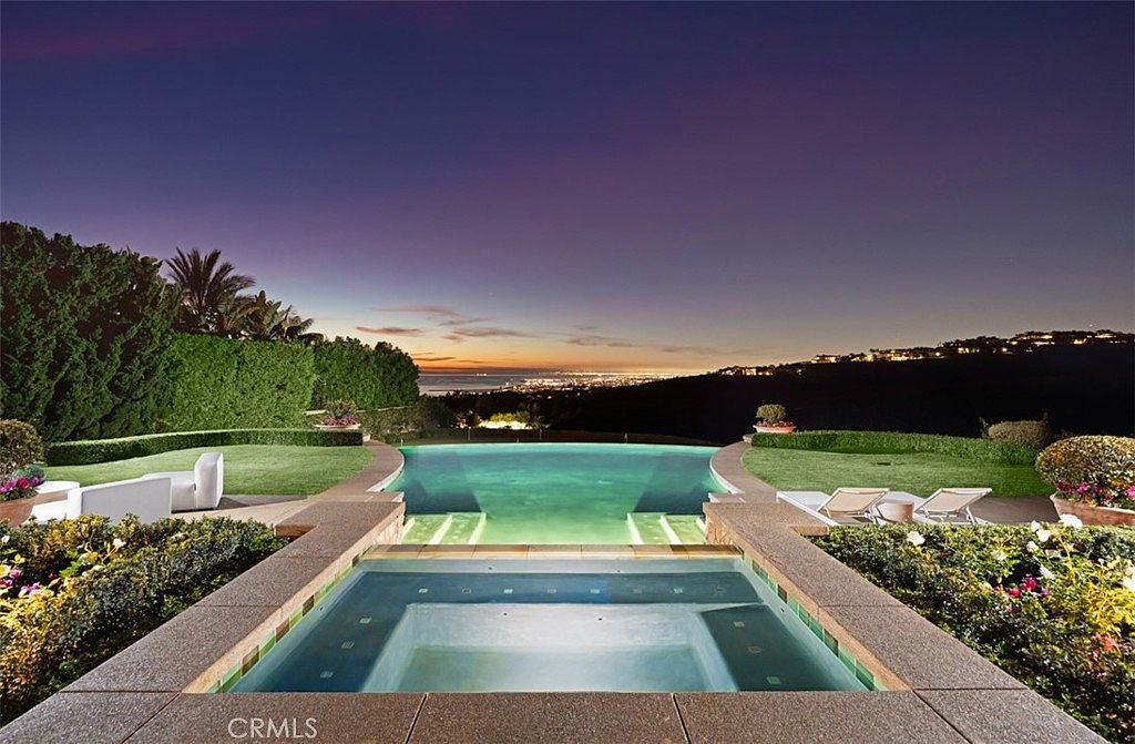 This image showcases a luxurious pool and spa area at dusk, featuring a multi-tiered design with illuminated steps leading into the pool. Lush landscaping surrounds the pool, and in the background, city lights twinkle against the horizon. The scene evokes a sense of tranquility and upscale living.