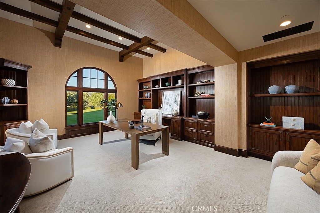 This is an interior shot of a well-appointed office or study. The room features dark wood built-in bookshelves, a large arched window overlooking a green lawn, and a substantial wooden desk. The room is decorated in neutral tones with textured wallpaper and includes comfortable seating, creating a sophisticated and inviting workspace.
