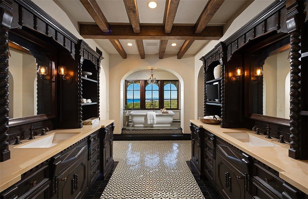 This is a luxurious primary bathroom featuring dual dark wood vanities with light countertops and integrated sinks. The room has a patterned tile floor and exposed wood beam ceiling. A freestanding bathtub is positioned in front of a large arched window offering an ocean view, creating a spa-like atmosphere.