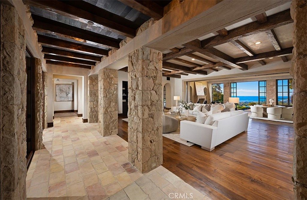 This interior shot showcases a luxurious living room with a seamless transition from a tiled hallway to hardwood floors. Stone pillars support a beamed ceiling, leading to a bright living area with white sofas and a stunning ocean view through large windows. The space exudes elegance and comfort, perfect for relaxation and entertaining.