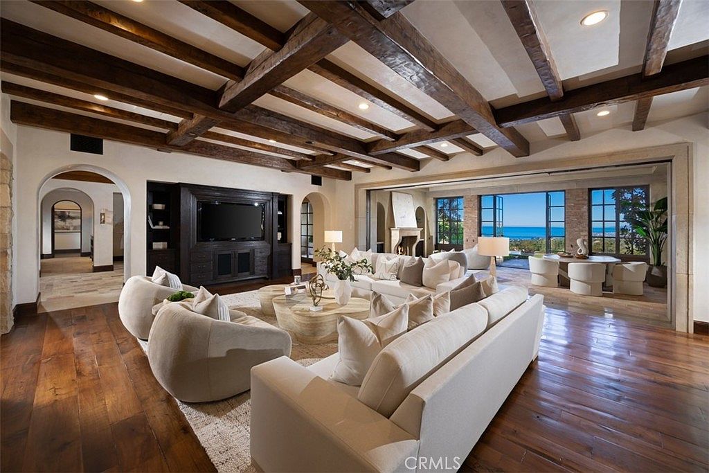 This is a spacious living room featuring hardwood floors, exposed wood beam ceilings, and a large built-in entertainment center. The room is furnished with comfortable white sofas and chairs, arranged around a light-colored area rug and coffee table. Large sliding glass doors offer a view of the ocean, creating a bright and airy atmosphere.