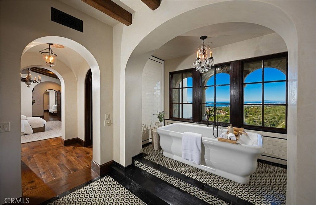 This is a luxurious primary bathroom featuring a freestanding white bathtub positioned in front of arched windows that offer a scenic view. The bathroom has a sophisticated design with black and white patterned tile flooring, arched doorways, and a decorative chandelier. The space exudes elegance and tranquility, making it a perfect retreat.