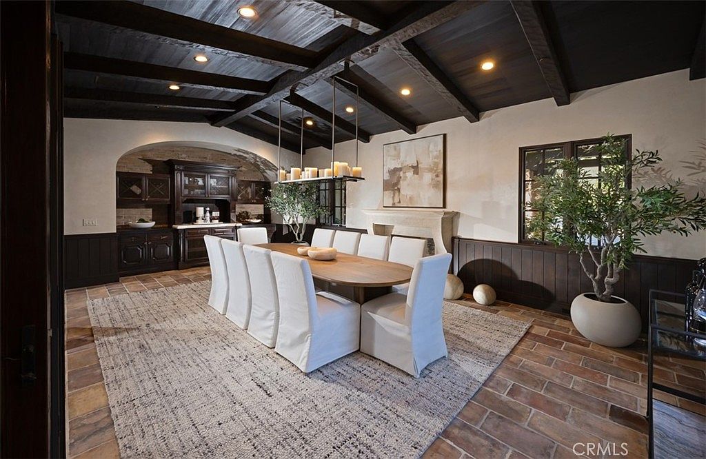 This is an interior shot of a dining room featuring a long oval wooden table surrounded by white upholstered chairs. The room has a rustic yet elegant feel, with dark wood ceiling beams, wainscoting, and brick flooring partially covered by a light-colored area rug. A large chandelier hangs above the table, and a decorative tree in a pot adds a touch of greenery.