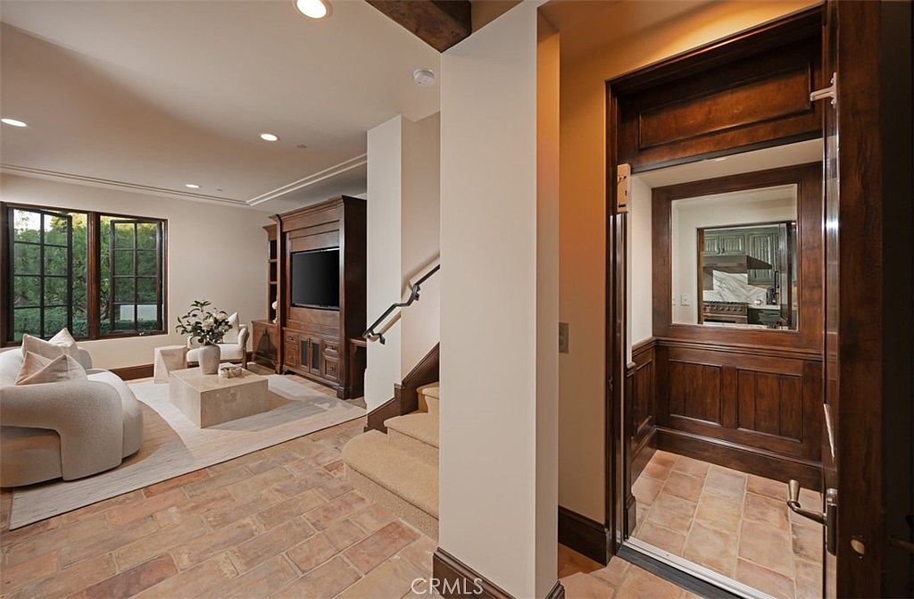This interior shot showcases a hallway with stairs and an elevator. The flooring is brick, and the walls are painted in a neutral tone. A living area is visible in the background, featuring a comfortable sofa and a built-in entertainment center, creating a seamless transition between spaces.