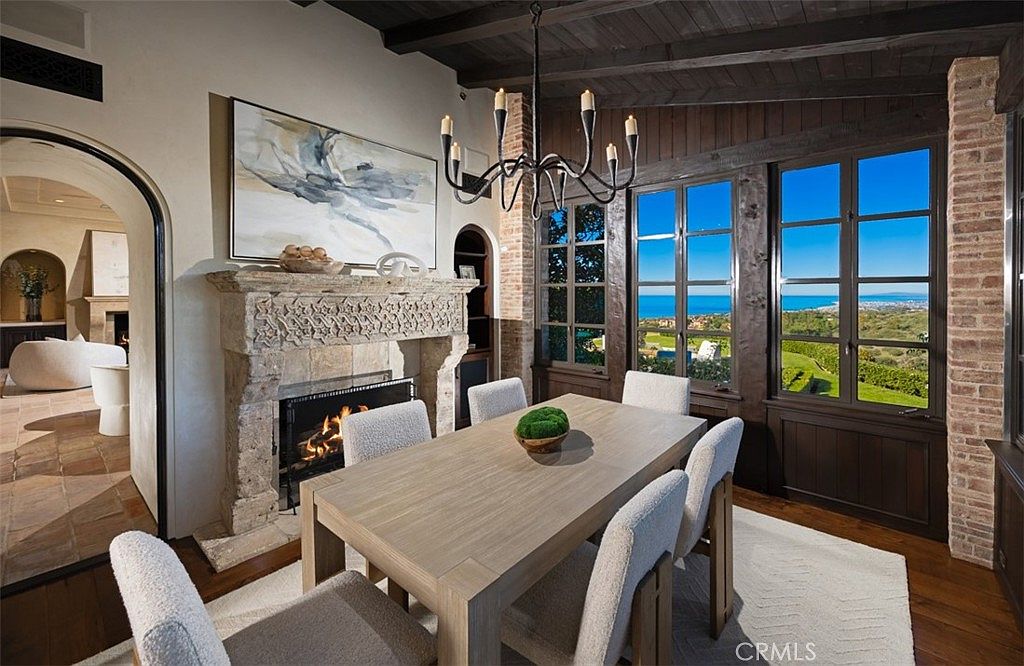 This is an interior shot of a dining room featuring a large wooden table with six chairs, a stone fireplace with a painting above it, and large windows offering a scenic view of the ocean and landscape. The room has a rustic yet elegant style, with exposed brick and wooden beams adding character. The overall impression is one of warmth and sophistication, perfect for entertaining or family meals.