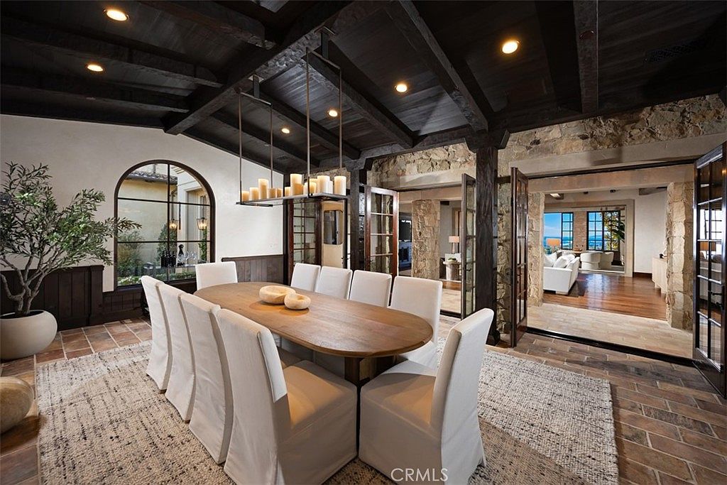 This is an interior shot of a dining room featuring a long, oval wooden table surrounded by white upholstered chairs. A modern chandelier hangs above the table, and the room has dark wood beams on the ceiling and stone accents on the walls. The space opens to another room with a view of the ocean, creating a luxurious and inviting atmosphere.