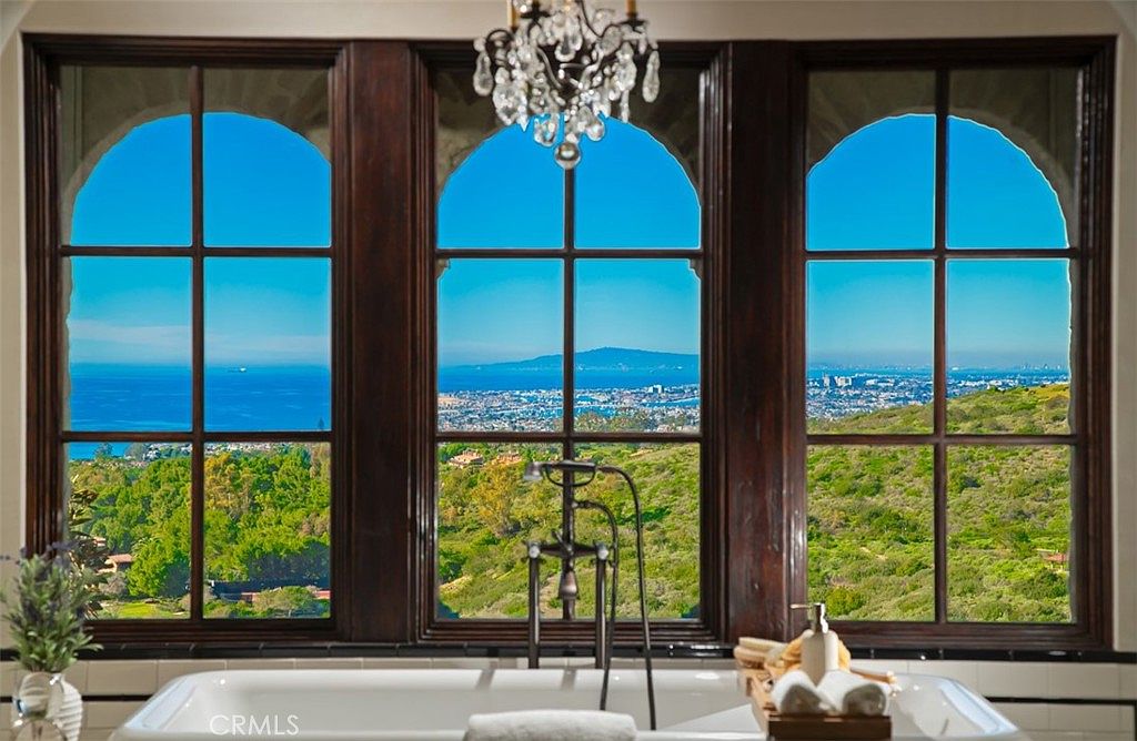 This image showcases a luxurious primary bathroom featuring a freestanding bathtub positioned in front of a large window offering a stunning view of the ocean and surrounding landscape. The bathroom is adorned with elegant fixtures, including a chandelier, and the window is framed by dark wood, adding a touch of sophistication. The overall impression is one of tranquility and opulence, perfect for relaxation.