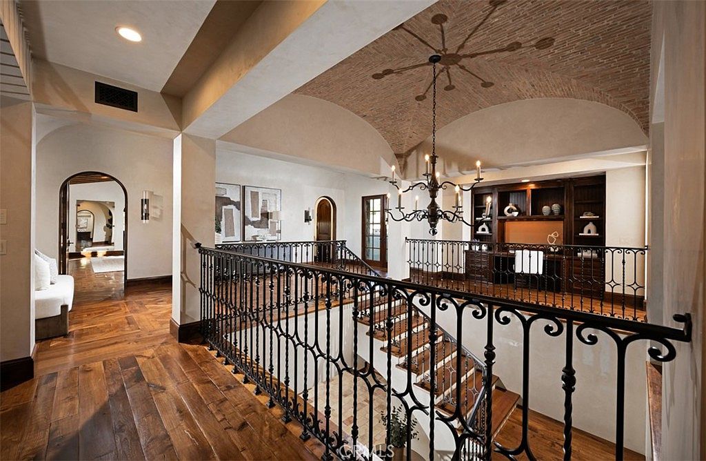 This interior shot showcases a grand hallway and staircase with wrought iron railings. The hallway features hardwood flooring, arched doorways, and decorative wall sconces. A barrel-vaulted ceiling with brickwork adds a touch of old-world charm, while a chandelier hangs elegantly above the staircase, creating a luxurious and inviting atmosphere.