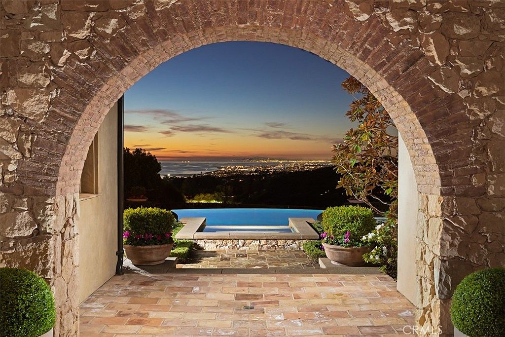 This image showcases a luxurious outdoor pool and spa area, framed by a rustic stone archway. The pool overlooks a stunning cityscape at dusk, creating a serene and picturesque setting. The brick-paved patio and manicured greenery add to the property's upscale appeal, emphasizing its potential for relaxation and entertainment.