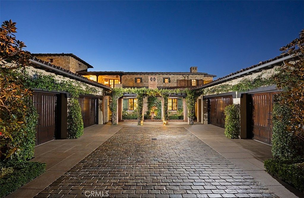 This is a front view of a luxurious estate at dusk, showcasing a grand courtyard paved with brick. The architecture features stone walls, dark wood garage doors adorned with climbing vines, and a pergola-covered walkway leading to the main entrance. The lighting highlights the texture of the stone and creates a warm, inviting atmosphere.