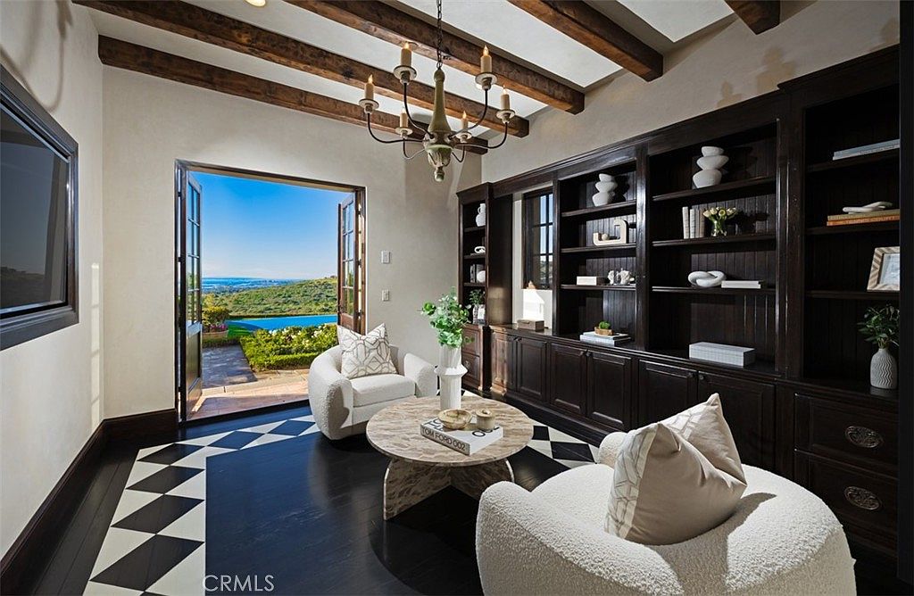 This interior shot showcases a sophisticated office or study with a dark wood built-in bookcase, two plush white armchairs, and a round marble-topped coffee table. The room features a black and white patterned floor and exposed wooden ceiling beams, adding character and warmth. French doors open to a stunning outdoor view, enhancing the room's appeal and creating a seamless indoor-outdoor connection.