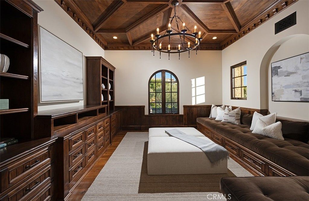 This is an interior shot of a luxurious office or study. The room features rich wood paneling, built-in shelving and cabinetry, and a coffered wood ceiling with a decorative chandelier. A long cushioned bench with pillows sits beneath a window, and a large ottoman is centered on a neutral rug, creating a sophisticated and comfortable workspace.