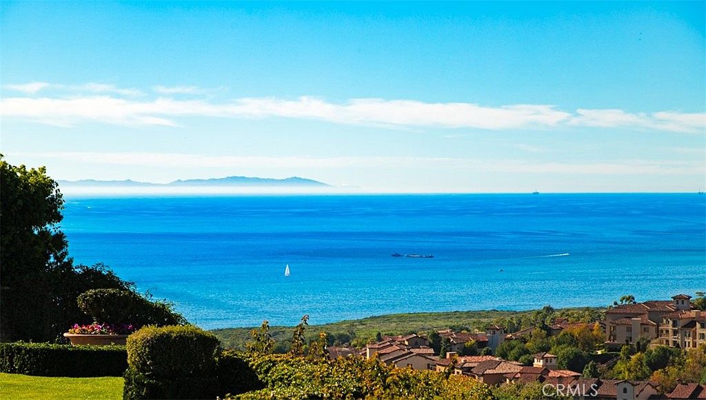 This aerial view showcases a stunning coastal property with a panoramic ocean vista. The foreground features lush greenery and terracotta-roofed buildings, leading to a vibrant blue ocean dotted with boats and a distant island on the horizon. The clear blue sky enhances the sense of tranquility and luxury, making it an ideal location for a high-end real estate listing.