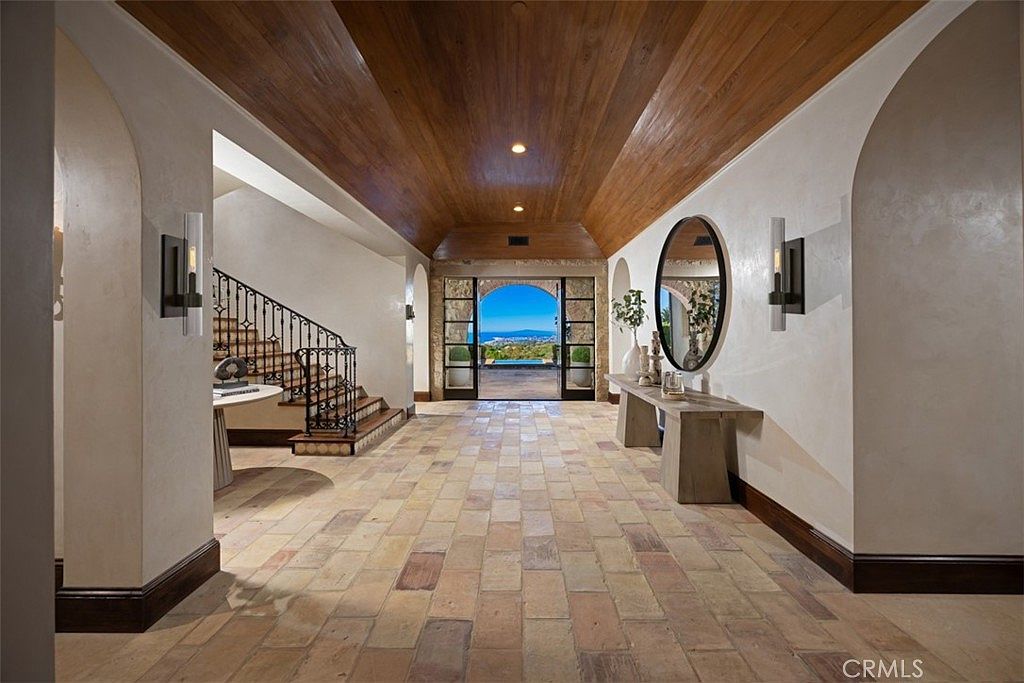This interior shot showcases a grand hallway with terracotta tile flooring and a wood-paneled ceiling. An arched doorway leads to an outdoor view, while a staircase is visible to the left. The space is decorated with sconces, a mirror, and a console table, creating an elegant and inviting entrance.