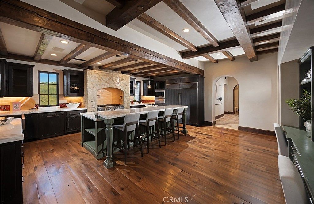 This is a spacious kitchen featuring dark wood cabinetry, a large central island with seating, and a stone-clad range hood. The ceiling is adorned with exposed wooden beams, adding a rustic touch to the space. Hardwood flooring extends throughout the kitchen, creating a warm and inviting atmosphere.
