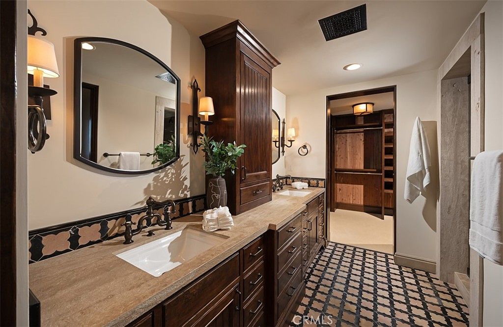 This is a primary bathroom featuring dark wood cabinetry with a light countertop and two sinks. A large mirror hangs above one sink, and sconces provide warm lighting. The floor is tiled in a decorative pattern, and a doorway leads to a walk-in closet.