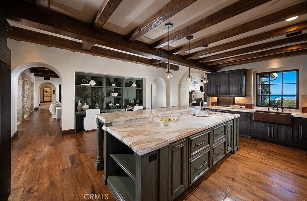 This is a spacious kitchen featuring a large island with a marble countertop, custom cabinetry, and wooden floors. The ceiling has exposed wooden beams and pendant lighting. A window provides a view of the outside, and there is an open hallway leading to other parts of the house.