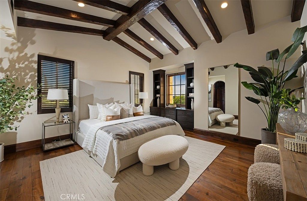 This is a primary bedroom featuring a vaulted ceiling with exposed wooden beams, creating a rustic yet elegant ambiance. The room is furnished with a large bed, a bench, and dark wood built-in cabinets flanking a window. A large mirror reflects the room, adding depth and light, while plants add a touch of nature.